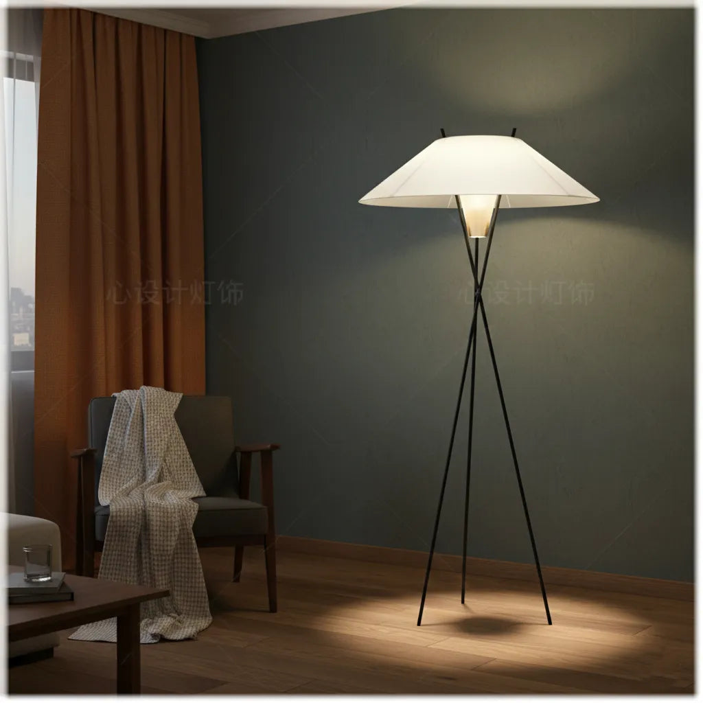 A modern floor lamp with a white conical shade and three black metal legs, illuminated in a dimly lit room with a dark gray wall, brown curtains, and a wooden chair.