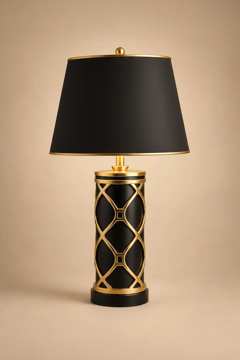 A table lamp with a black fabric drum shade, a black cylindrical base featuring a gold geometric lattice pattern, and gold metal trim, set against a solid beige background.