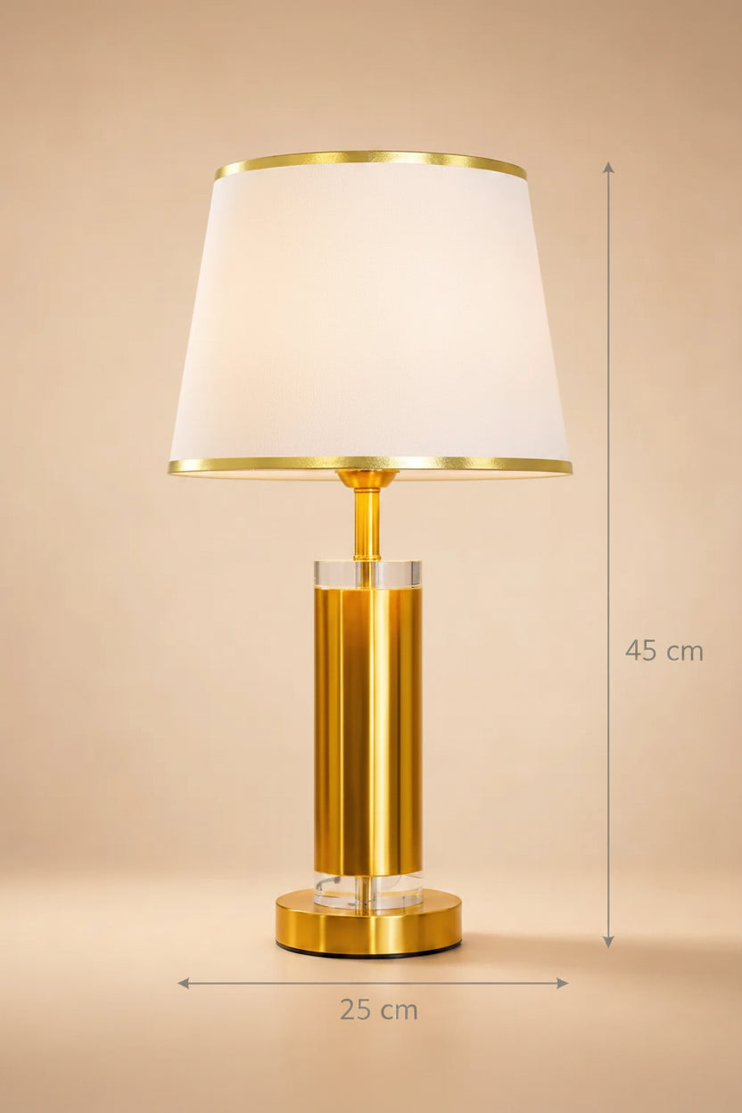 A modern table lamp with a white tapered fabric shade, a brushed gold metal base, and clear crystal accents, set against a warm beige background.