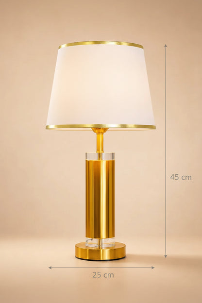 A modern table lamp with a white tapered fabric shade, a brushed gold metal base, and clear crystal accents, set against a warm beige background.