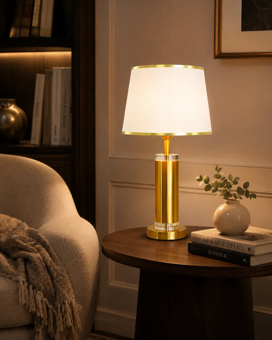 A modern table lamp with a glowing white drum shade and a polished gold cylindrical base, styled on a dark wood side table next to a beige armchair in a cozy room.