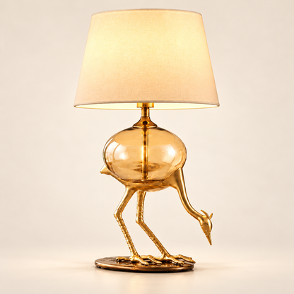 Aurelia gold-tone crane table lamp with amber glass body on a clean warm-white background